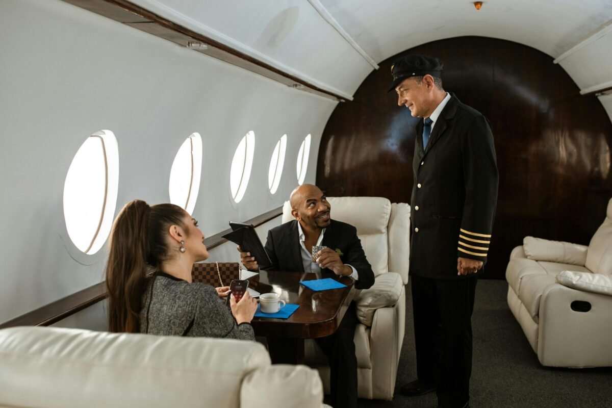 alt= Two passengers seated at a table in a private jet, having a conversation with the pilot who is standing and smiling. The passengers are holding a tablet and coffee cups are on the table.