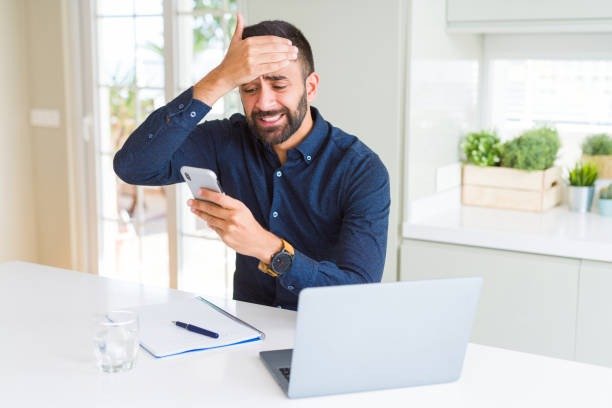 Sorry, unexpected news! A distressed man holds his smartphone while sitting at a desk with a laptop, reacting with frustration.